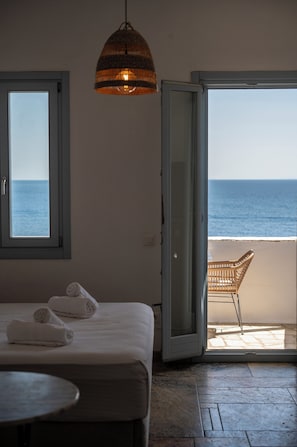 A room with a bed, a table, a hanging light fixture, a chair, and a view of the sea through an open door.