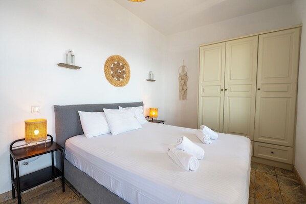 A neatly made bed with white linens, a bedside lamp, and a wall-mounted decorative item. - Akti Giannaki (Tinos)