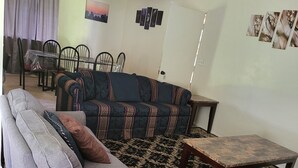 A living room with a sofa, a coffee table, a dining table with chairs, a ceiling fan, and a patterned rug.