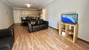 A modern living room with a black leather sofa, wooden flooring, a television on a wooden stand, and a kitchen in the background. - 115 AN - Stay Napa Oniro Apartment (Ayia Napa)