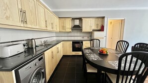 A compact kitchen with wooden cabinets, a washing machine, a toaster, and a dining table with chairs. - 115 AN - Stay Napa Oniro Apartment (Ayia Napa)