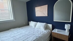 Un dormitorio con una cama, una mesita de noche, un espejo en la pared y una ventana con persianas.