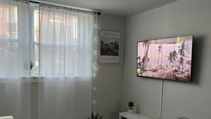 Una sala moderna con un televisor de pantalla plana, un sofá blanco, una mesa de centro con jarrones, una estantería y una ventana con cortinas ligeras.