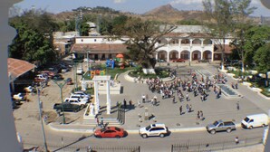 Una piazza con una fontana al centro, circondata da un edificio bianco con archi. Auto parcheggiate attorno alla piazza e alcune persone che passeggiano.