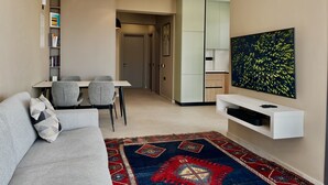 A modern living room with a sofa, a dining table with chairs, a TV mounted on the wall, and a kitchen area in the background. - 107 LIM - Stay Mouson View Apartment (Limassol)