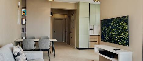 A modern living room with a sofa, a dining table with chairs, a TV mounted on the wall, and a kitchen area in the background.