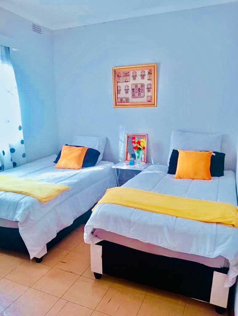 A room with two beds, each with a pillow and a blanket, a small table with a vase, and a framed picture on the wall.