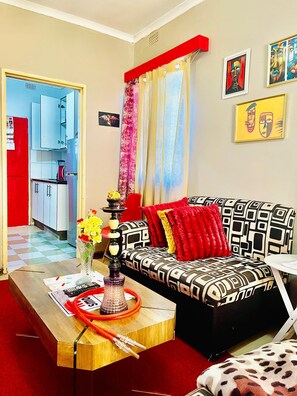 Un salon avec un canapé à motifs, une table basse sur laquelle se trouve un narguilé, une porte rouge et une cuisine visible à travers une porte ouverte.