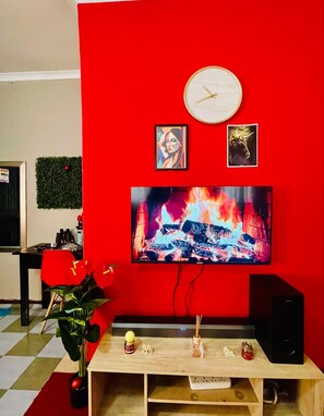 A living room with a red wall, a flat-screen TV, a clock, and a small table with decorative items. - 2-bedroom Family/Modern Apartment with City Views Pleasant  apartment in Mbabane (Mbabane)