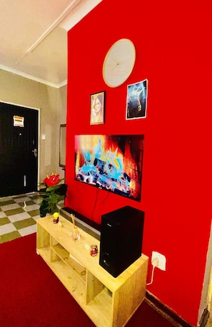 Une pièce avec un mur rouge, un téléviseur fixé au mur, un banc en bois et une porte au fond.