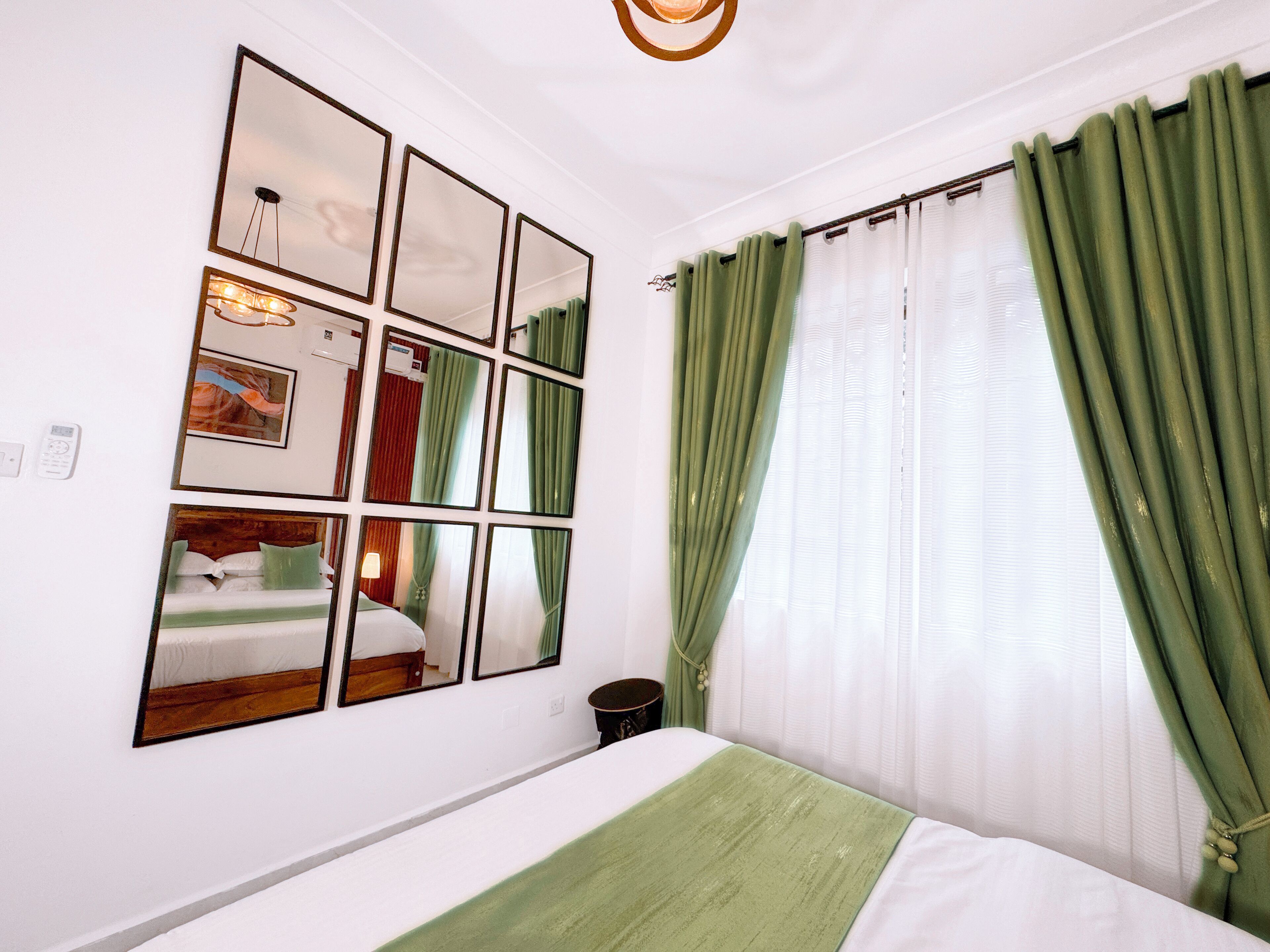 Una camera d'albergo con un grande specchio, un letto con una coperta verde, tende bianche e una tenda verde alla finestra.