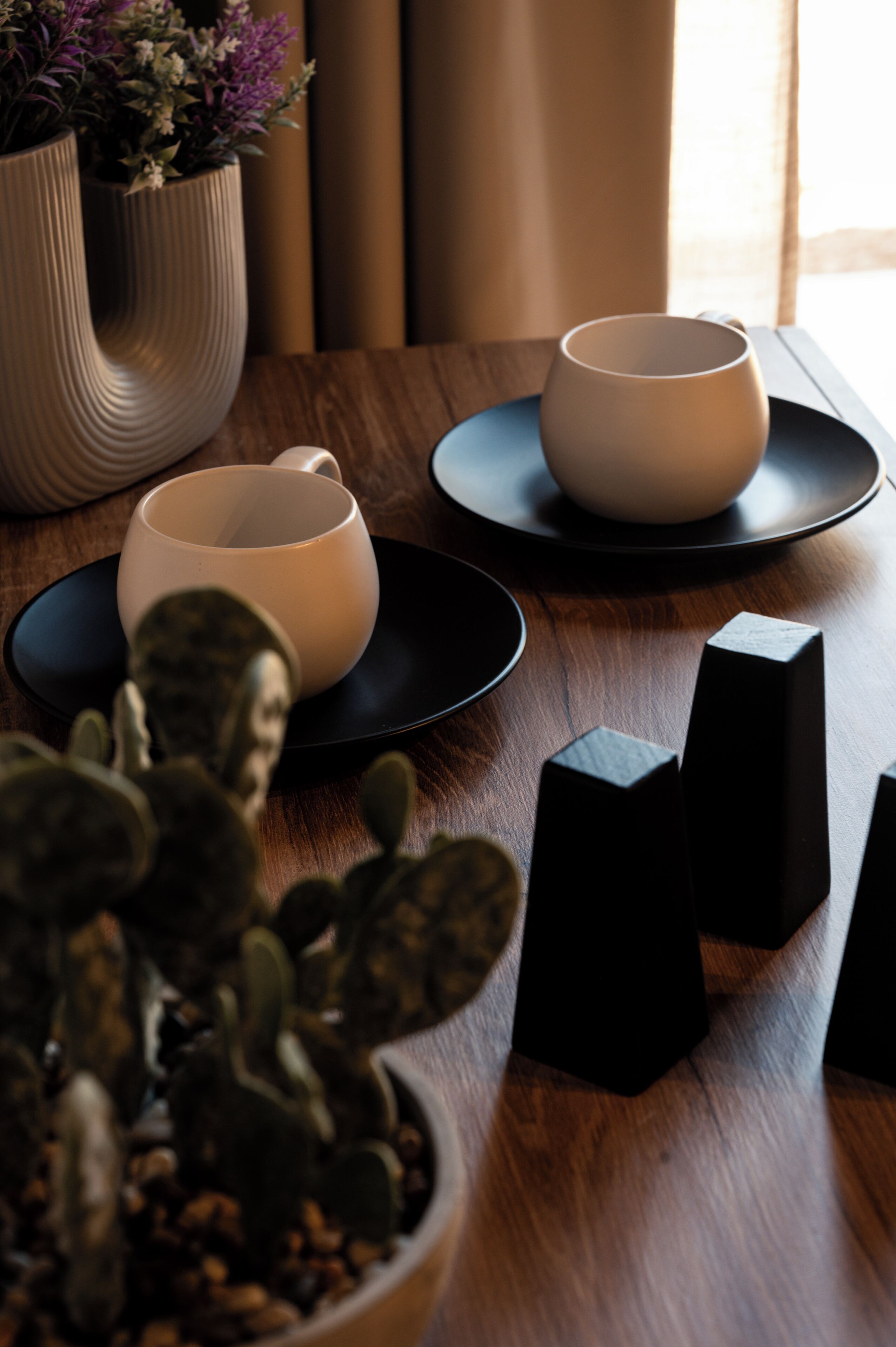 A wooden table with two white cups on black saucers, a potted plant, and black candle holders.