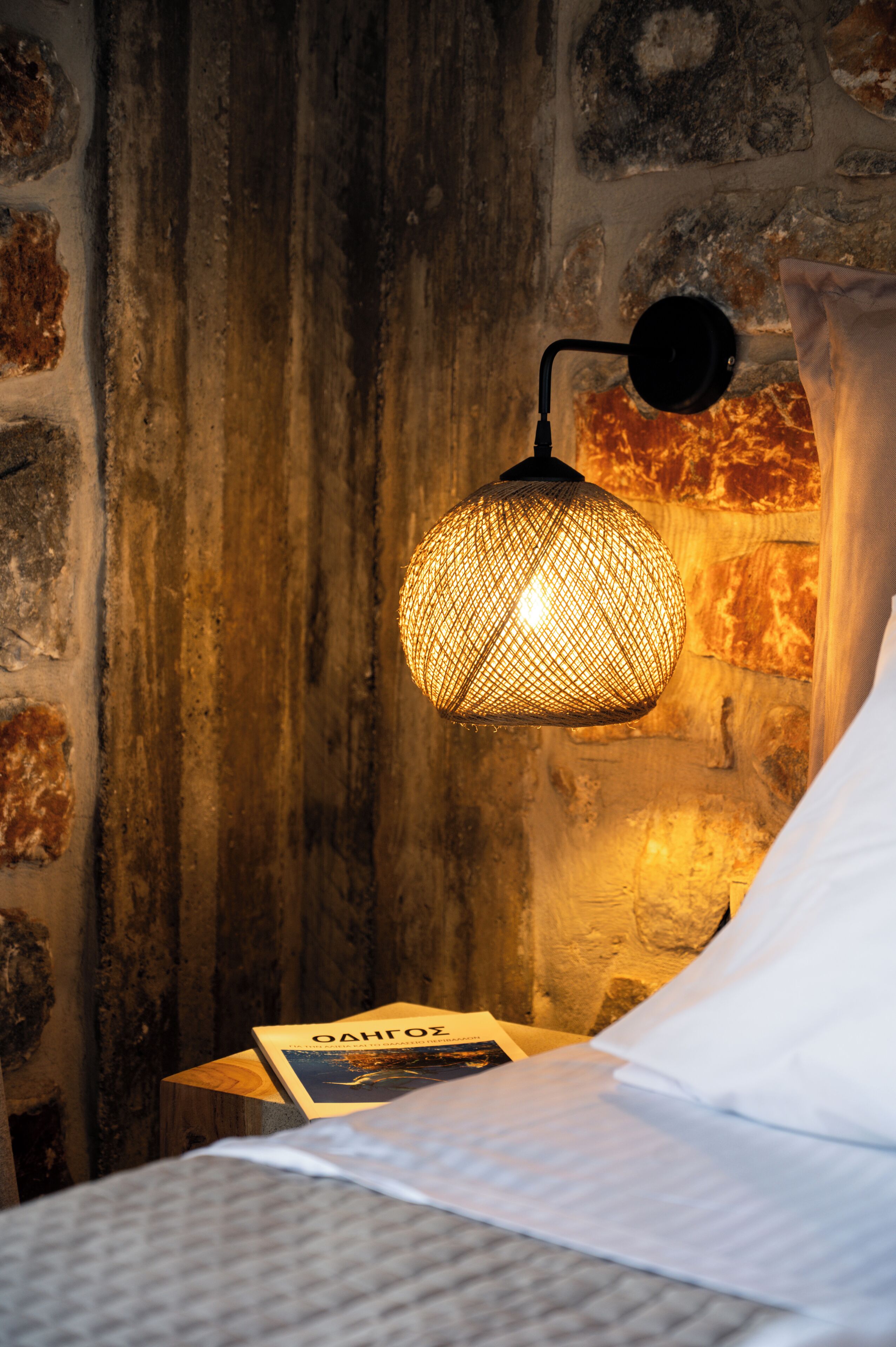 Ein Bett mit einem weißen Kopfkissen und einem Buch mit dem Titel "OAHUROZ" auf einem hölzernen Nachttisch. Eine gewebte Pendelleuchte hängt über dem Bett und beleuchtet die rustikale Steinwand dahinter.