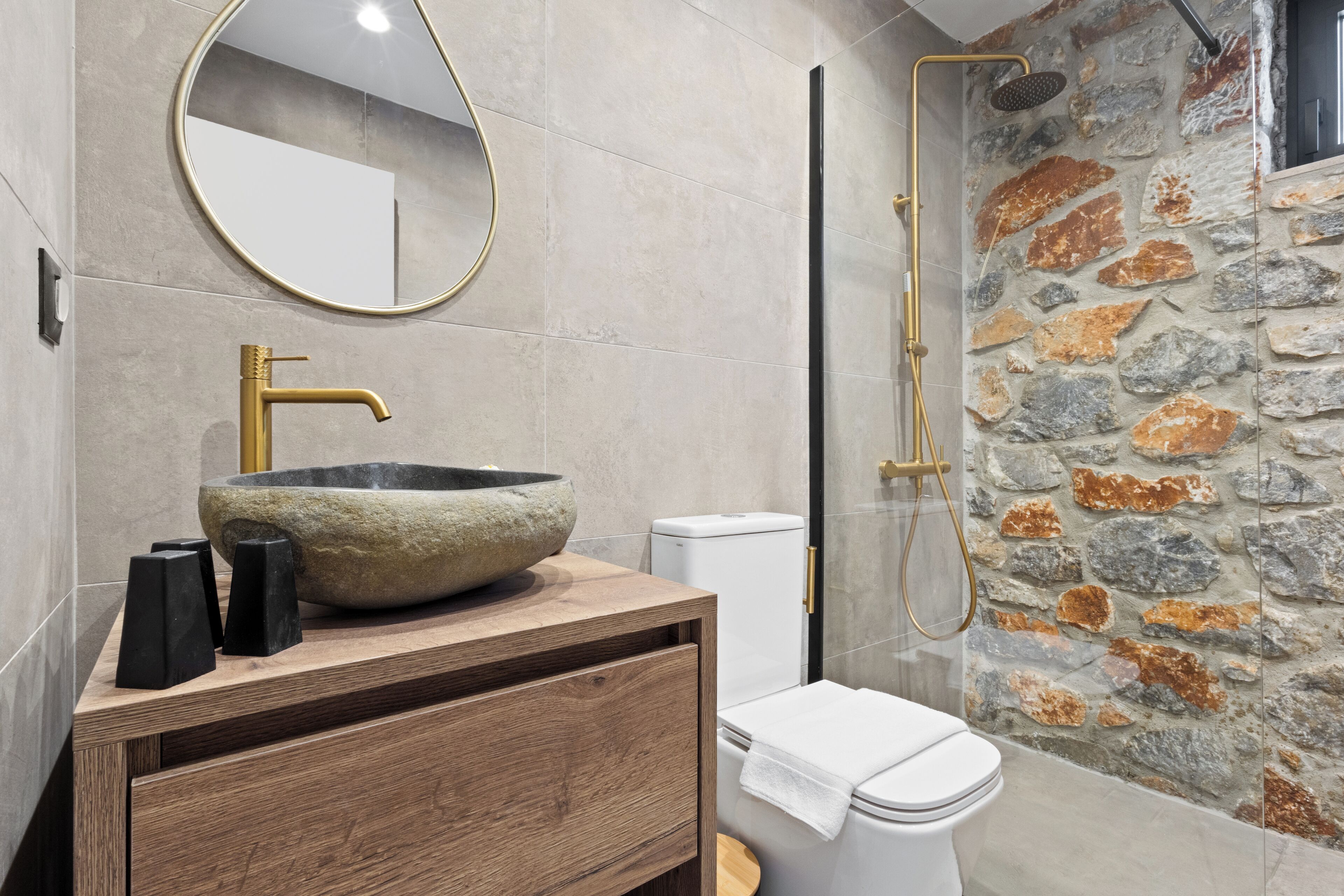 A bathroom with a stone sink, a wooden vanity, a toilet, and a shower with a stone wall.