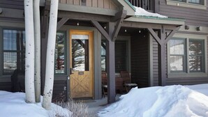 Une maison en bois avec un porche à l’avant, une porte portant l’inscription « 102 », et un sol recouvert de neige.