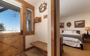 A room with a wooden door, a bench, a bed, and a view of a snowy landscape.