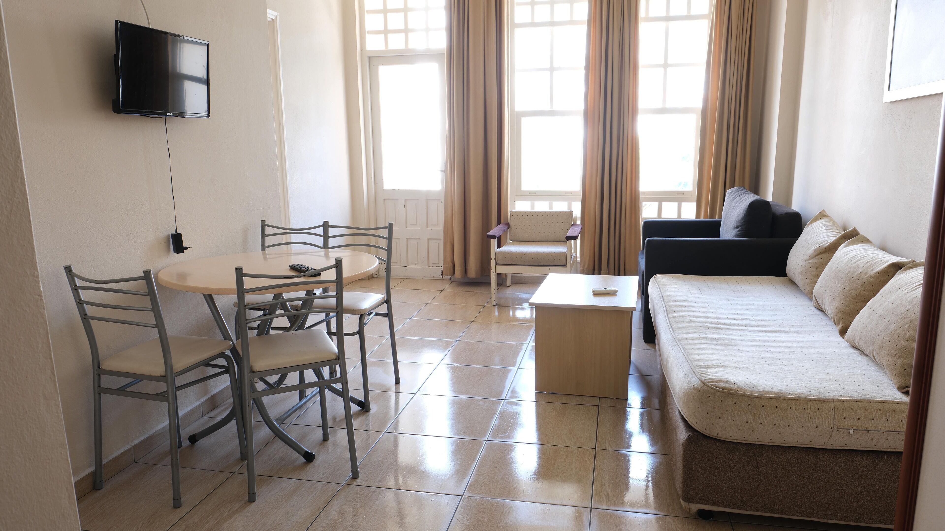 Una sala de estar con un sofá, una mesa de comedor con sillas, un televisor montado en la pared y ventanas amplias con cortinas.