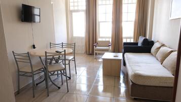 Una sala de estar con un sofá, una mesa de comedor con sillas, un televisor montado en la pared y ventanas amplias con cortinas.