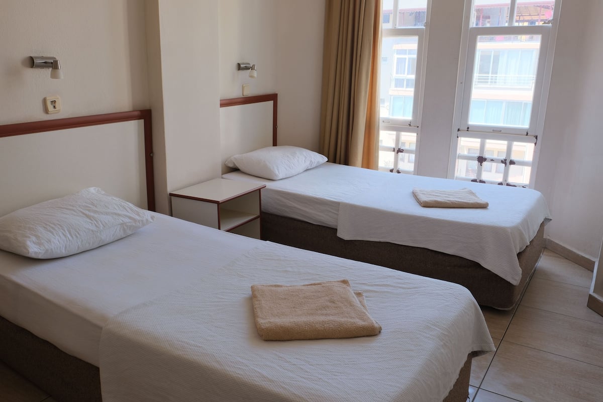 Habitación de hotel con dos camas individuales, cada una con una almohada y una toalla doblada. Hay una mesita de noche pequeña entre las camas. La habitación tiene una ventana con cortinas y paredes blancas.