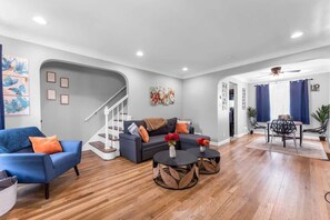 A modern living room with a grey sofa, blue armchair, and two round coffee tables with leaf-patterned bases. A staircase with a white railing leads to a second floor. - Brooklyn Townhome| Updated Kitchen| Sleeps 9 (Brooklyn)
