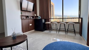 A modern living room with a flat-screen TV mounted on a wooden wall, a black refrigerator, a round wooden table with a vase, and a blue carpet. - Batroun Staycation (Batroun)