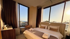 A hotel room with a large bed, a desk, a chair, and a view of the cityscape through large windows. - Batroun Staycation (Batroun)