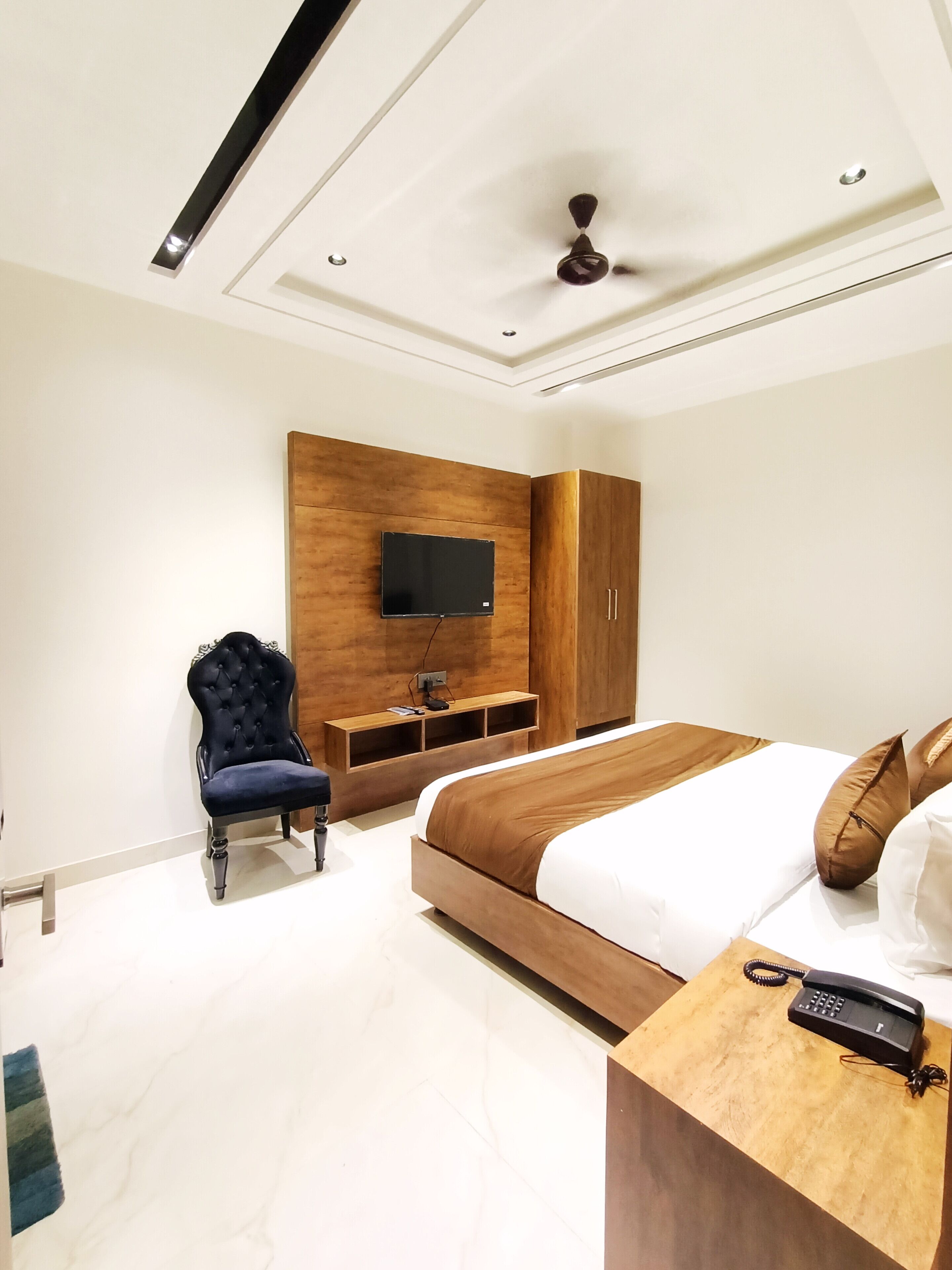 Un dormitorio moderno con una cama grande, un televisor montado en un panel de madera, un sillón tapizado oscuro y una mesita de noche de madera con un teléfono.