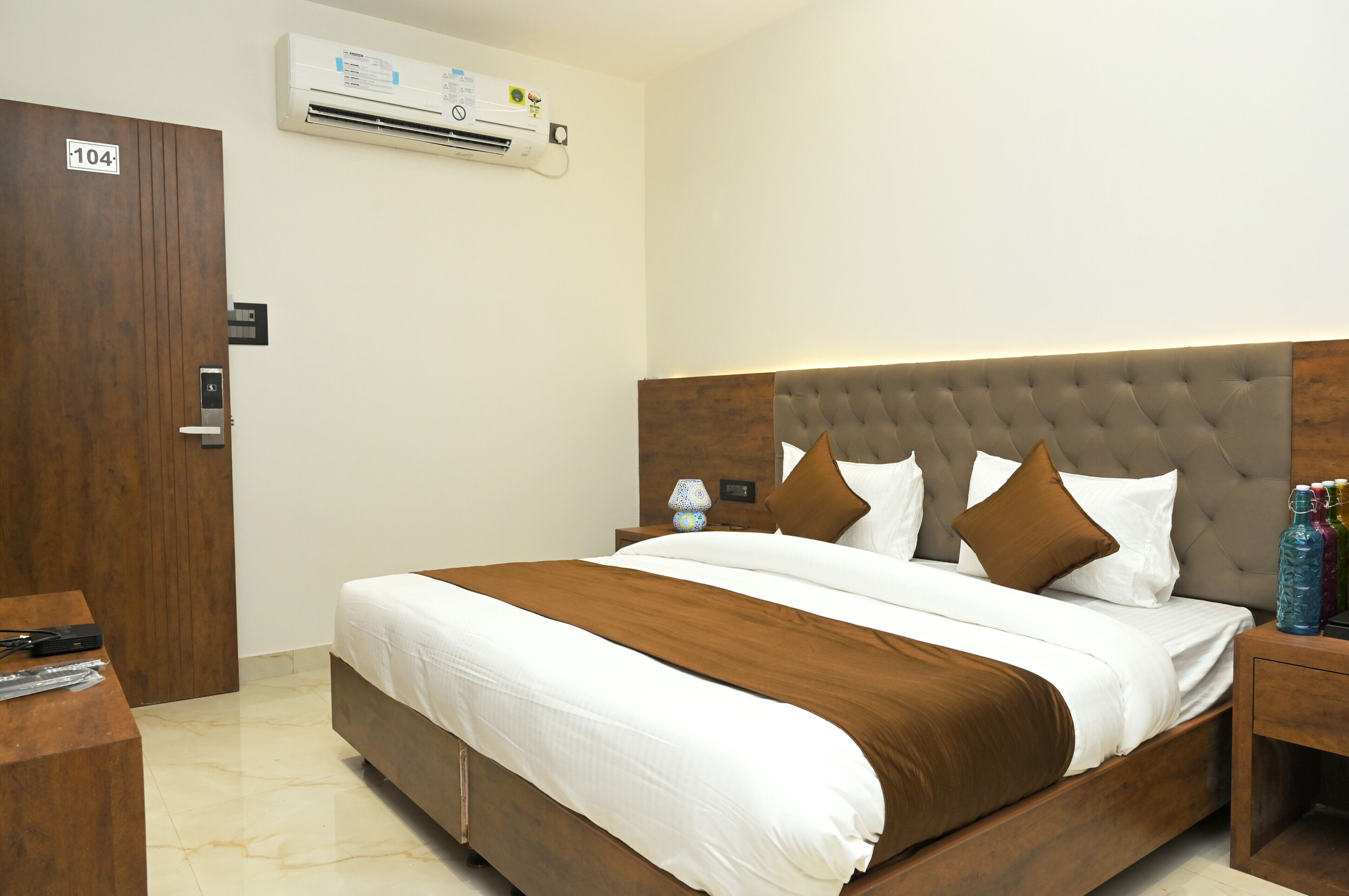 Una habitación de hotel con una cama grande, un cabecero de madera, una mesita de noche con lámpara y un equipo de aire acondicionado en la pared.