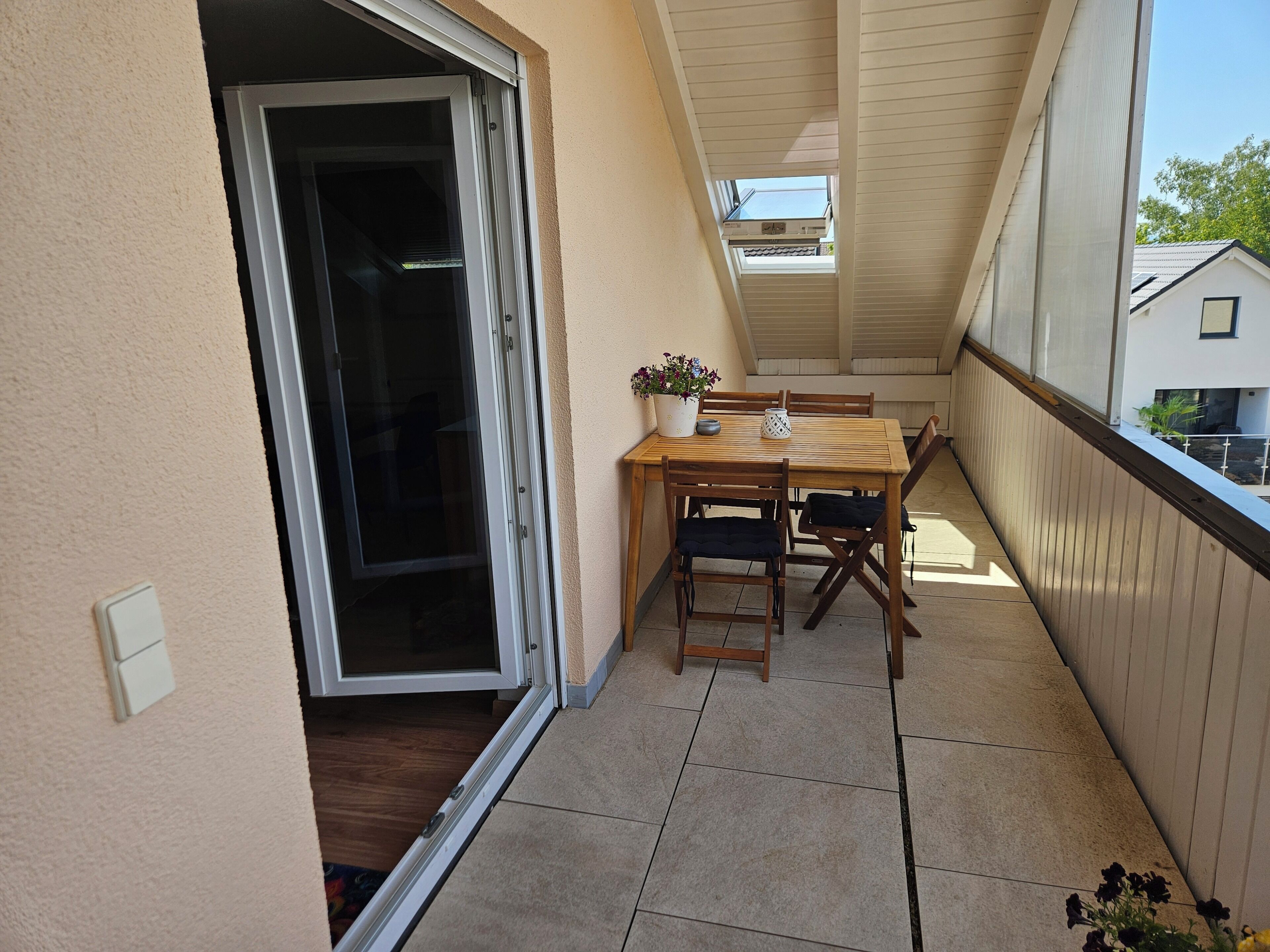 Un balcón con una mesa y sillas de madera, una planta en maceta y una puerta corrediza de vidrio que da a un espacio interior.