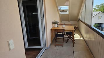 Un balcón con una mesa y sillas de madera, una planta en maceta y una puerta corrediza de vidrio que da a un espacio interior.