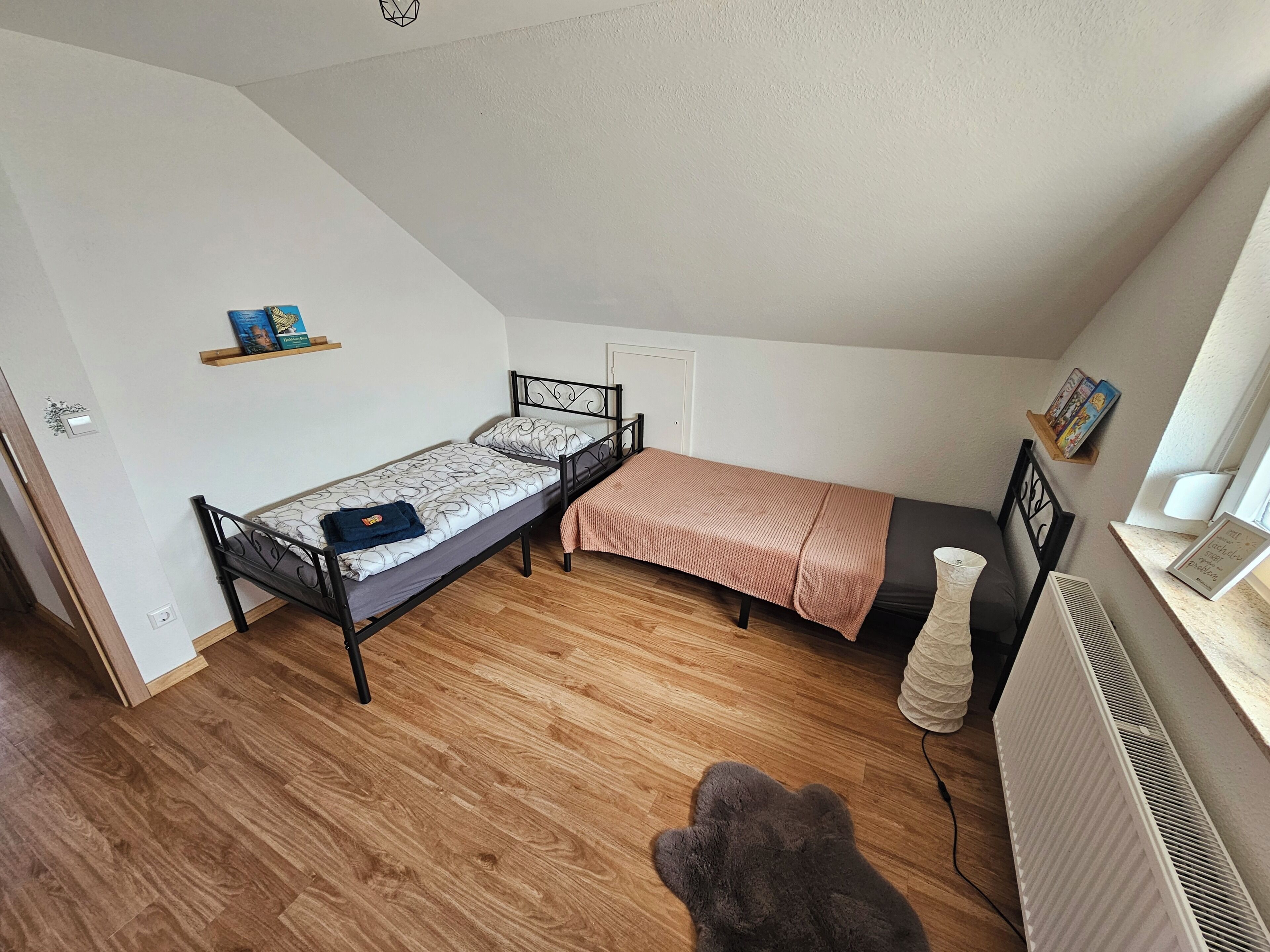 Una habitación pequeña con cama individual, techo inclinado, un radiador y una estantería con libros.