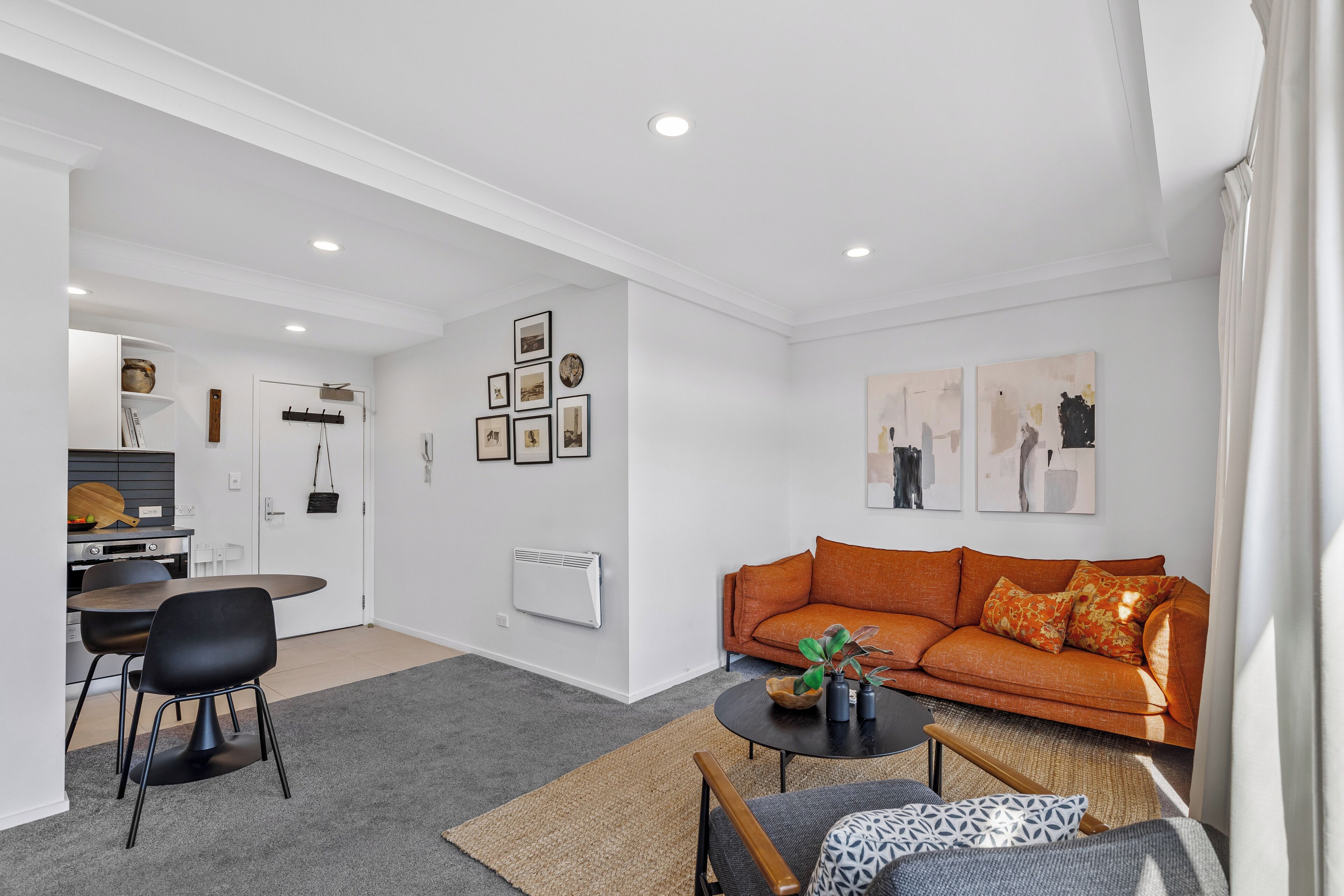 Een moderne woonkamer met een bank, salontafel en stoelen. Er is een eethoek met een tafel en stoelen. De kamer heeft witte muren en een plafond, en er hangen foto's aan de muur.