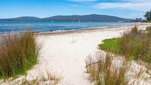 En strand med sand, gräs och utsikt över havet och avlägsna kullar.