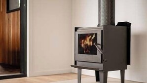 En modern vedeldad kamin med en glaslucka, placerad i ett rum med trägolv och en vit vägg.