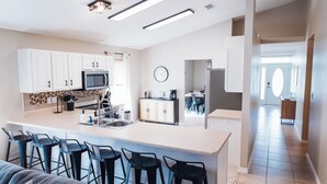 A modern kitchen with white cabinets, a central island with bar stools, a microwave, and a clock on the wall. - Golden Hour Getaway-2 Minutes to Beach, Pool + Arcade, Sleeps 10, (Destin)