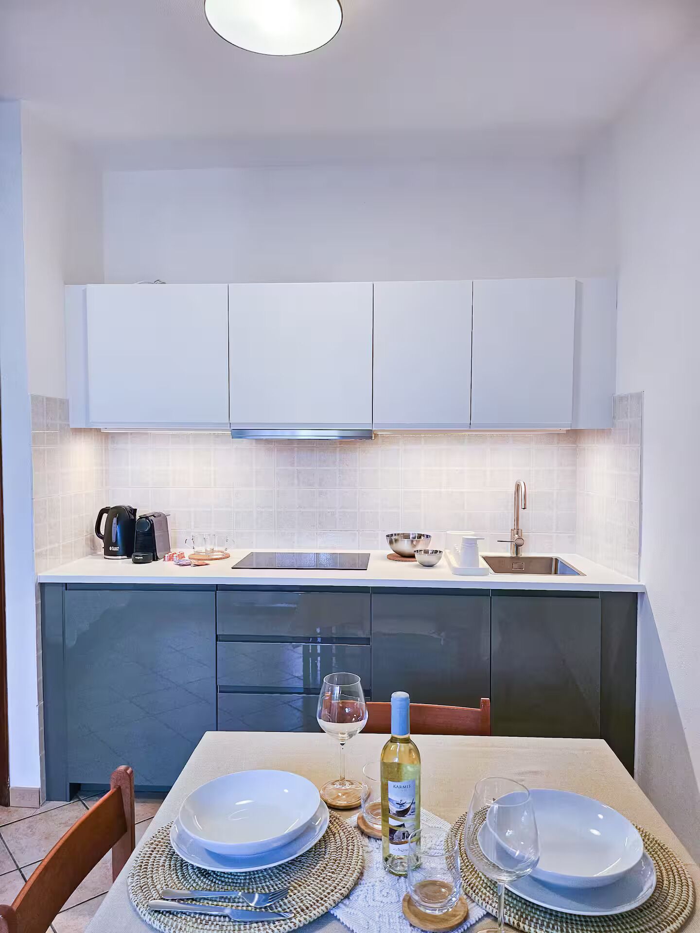 Una cocina moderna con armarios blancos, un lavamanos de acero inoxidable y una mesa de comedor dispuesta con platos, vasos y una botella de vino.