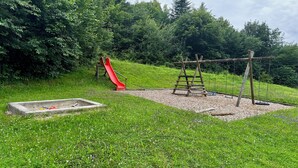 Une aire de jeux avec un toboggan rouge et des balançoires, située dans un espace verdoyant avec des arbres en arrière-plan.