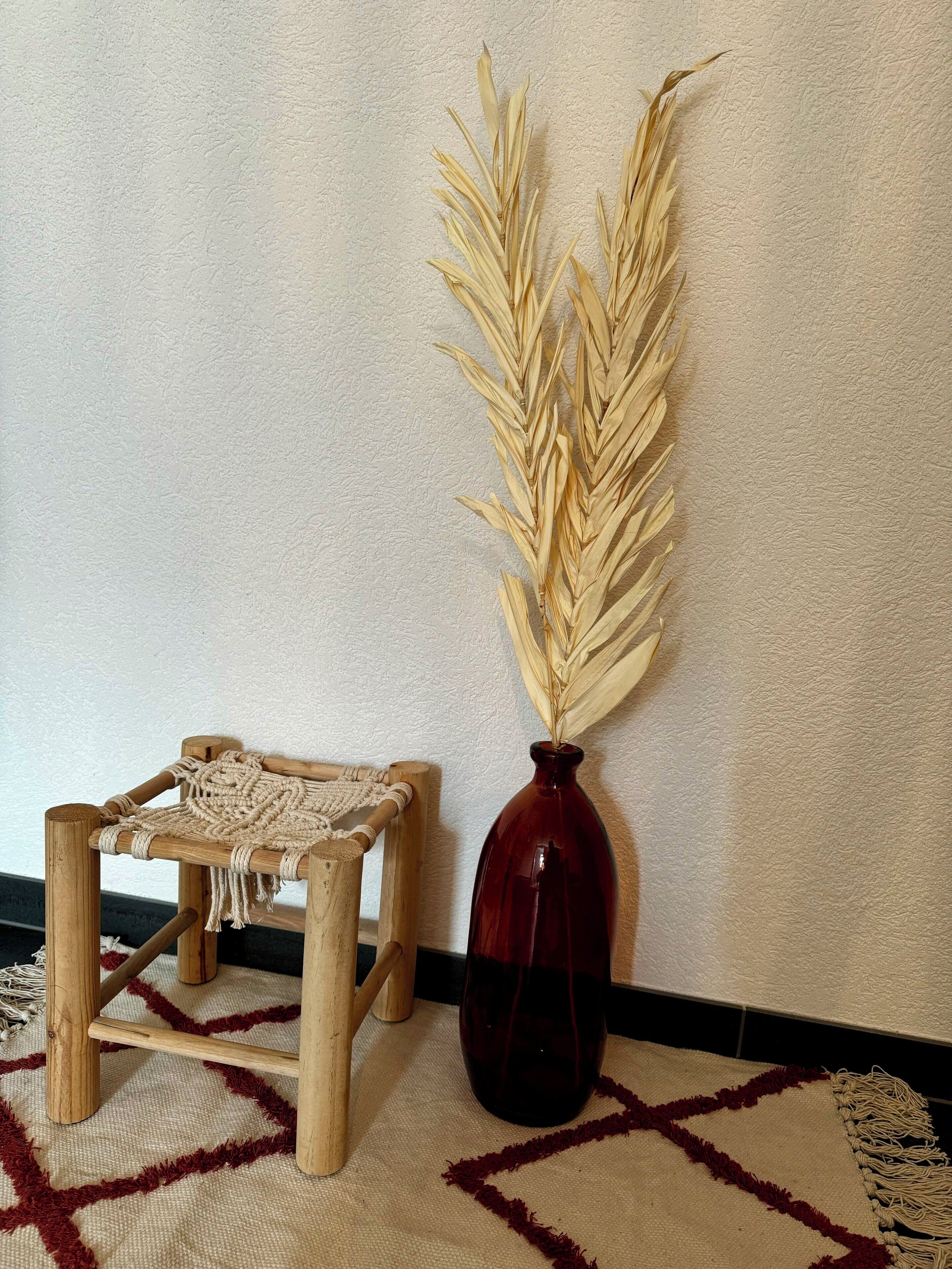 A small wooden stool with a macrame seat, a dark red vase with dried wheat stalks, and a geometric patterned rug on the floor.