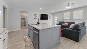 A modern kitchen with granite countertops, a built-in dishwasher, and a central island. There is a gray sofa with red and white pillows, a flat-screen TV mounted on the wall, and a ceiling fan. - Sporty Spot - Hot Tub - Walk to AT&T Stadium (Arlington)