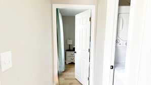 En hall som leder till ett sovrum med en vit dörr, ett nattduksbord, en lampa och en tvättmaskin.