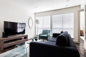 Un salón moderno con un televisor de pantalla plana, una mesa de centro de cristal, un sofá oscuro y un sillón azul claro.