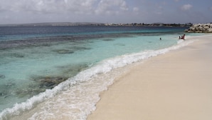 Ein Strand mit klarem türkisblauem Wasser, einem Sandufer und einer entfernten Küstenlinie mit Gebäuden.