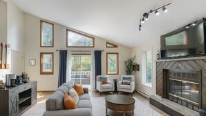 Ein geräumiges Wohnzimmer mit grauem Ecksofa, zwei Sesseln, einem Couchtisch, einem Kamin und großen Fenstern.