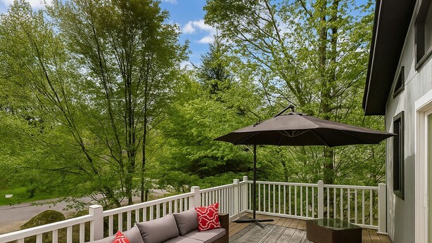 Eine Terrasse mit einer Couch und einem Sonnenschirm, umgeben von weißen Geländern und viel Grün.