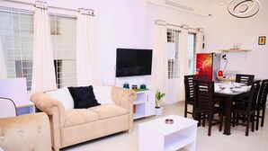 Una sala de estar con un sofá color beige, una mesa de centro blanca, una mesa de comedor con sillas y un televisor de pantalla plana montado en la pared.