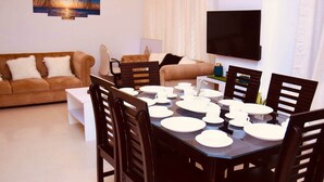 Comedor con una mesa dispuesta para seis personas, rodeada de sillas de madera oscura. Se ve un sofá y un televisor al fondo.