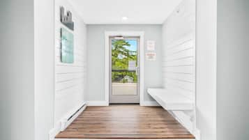 A narrow hallway with white walls and wooden flooring, leading to a door with a view of a green landscape.