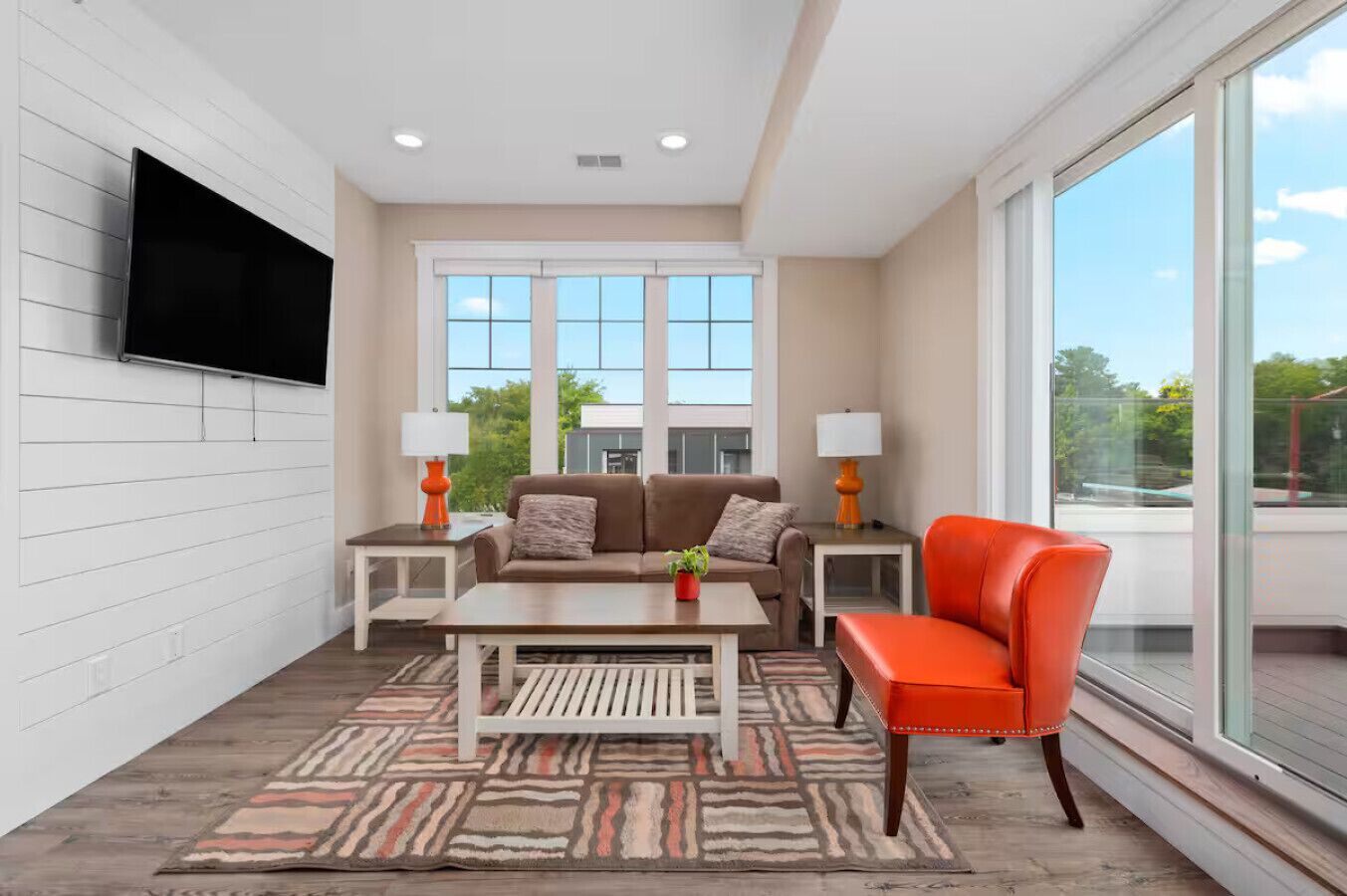 A modern living room with a sofa, coffee table, and a bright orange chair. There are two side tables with lamps, a flat-screen TV mounted on the wall, and large windows offering a view of the outdoors.