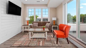 Una sala moderna con un sofá, una mesa de centro y un sillón naranja brillante. Hay dos mesas auxiliares con lámparas, un televisor de pantalla plana montado en la pared y ventanas amplias que ofrecen vistas al exterior.