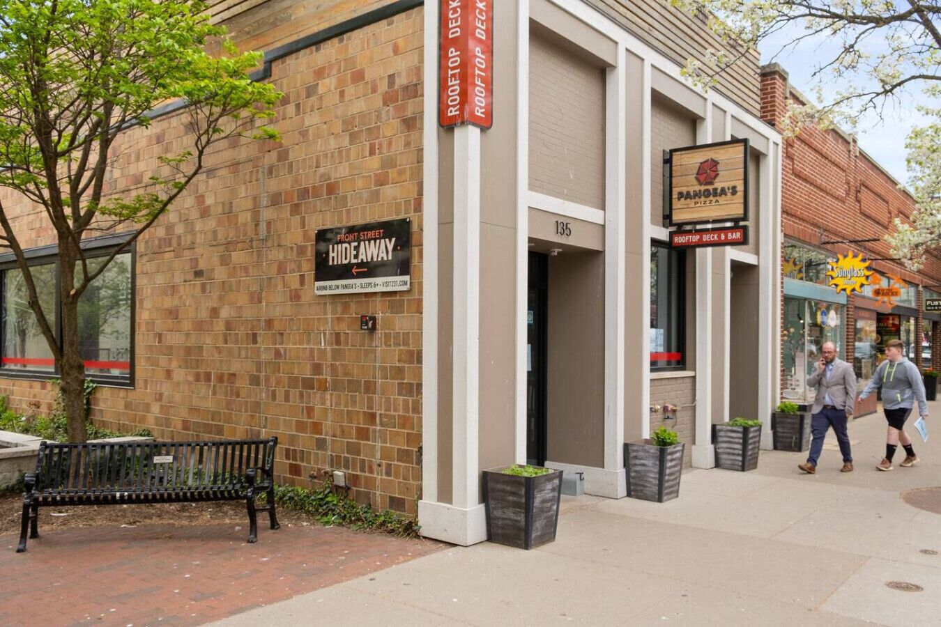 A brick building with a sign for 'Rooftop Deck & Bar' and 'Panda's Pizza'. There is a bench and potted plants on the sidewalk.